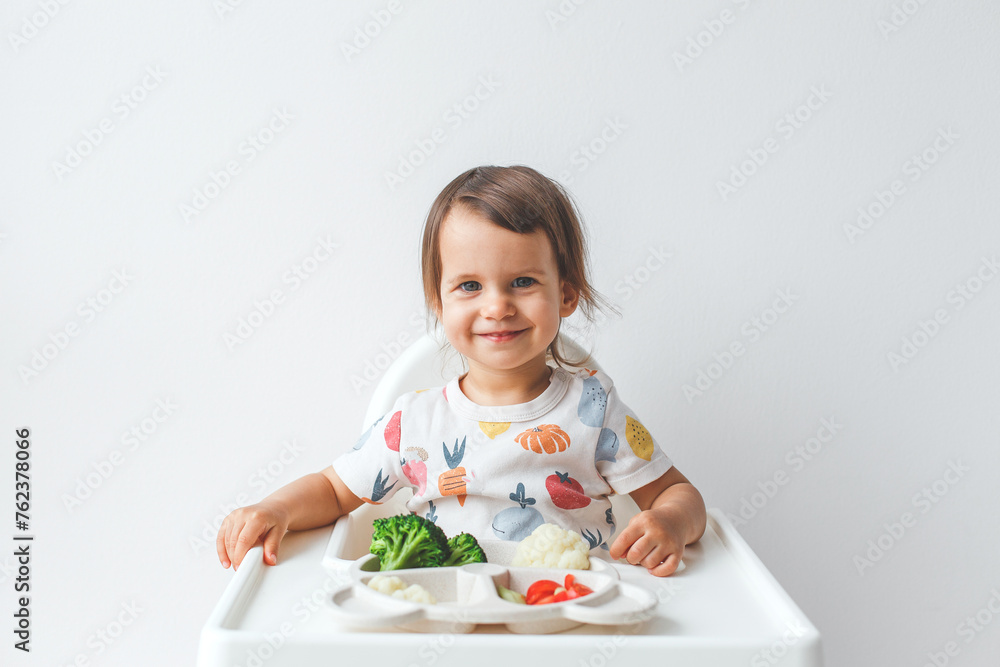 a happy 1.5 year old child sits in a high chair on a white background with a plate of vegetables, smiles and looks at the camera, concept of baby food and complementary feeding a happy 1.5 year old child sits in a high chair on a white background with a plate of vegetables, smiles and looks at the camera, concept of baby food and complementary feeding