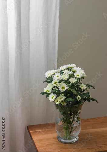 A bouquet of white chrysanthemums in a glass vase on a wooden bench in the living room