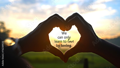 Inspirational quote - We can only learn to love by loving. On person making heart shape for love sign with fingers against the colorful sunset sunrise light background. Romantic love quotes concept.