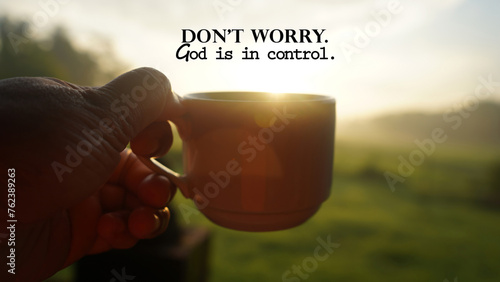Morning inspirational quote - Don't worry. God is in control. With person holding a cup of coffee or tea against the warm sunrise light background. Faith and hope. Believe in God concept. photo