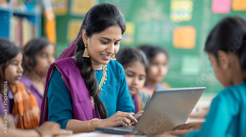 A young Indian lady teacher conducts an online class on a laptop in a government school classroom, surrounded by students in uniform