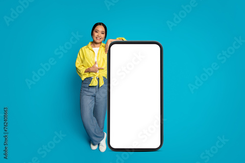 Cheerful woman in a yellow blouse and jeans leaning on a large vertical smartphone with a blank screen