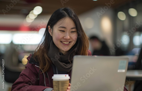 An Asian woman smiling while using her laptop at a cafe table with a coffee cup