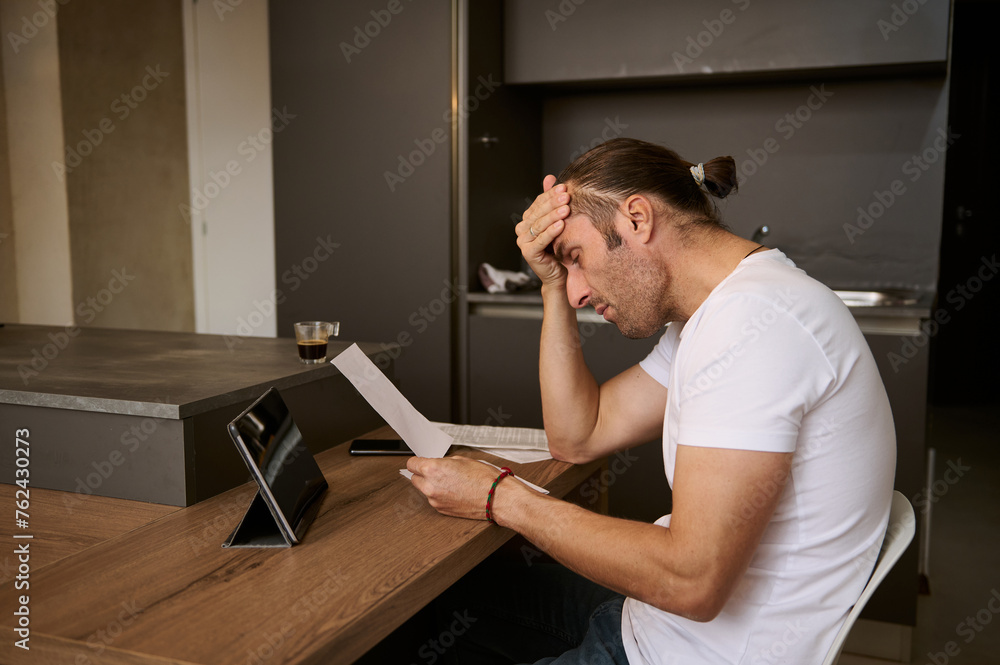 Young man sitting at the table, putting his hand on his forehead, feeling frustration and shock by costs of paying bills Young man sitting at the table, putting his hand on his forehead, feeling frustration and shock by costs of paying bills