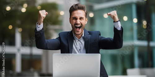 A young businessman celebrating success with arms raised in front of a laptop, looking a laptop in a happy and successful pose