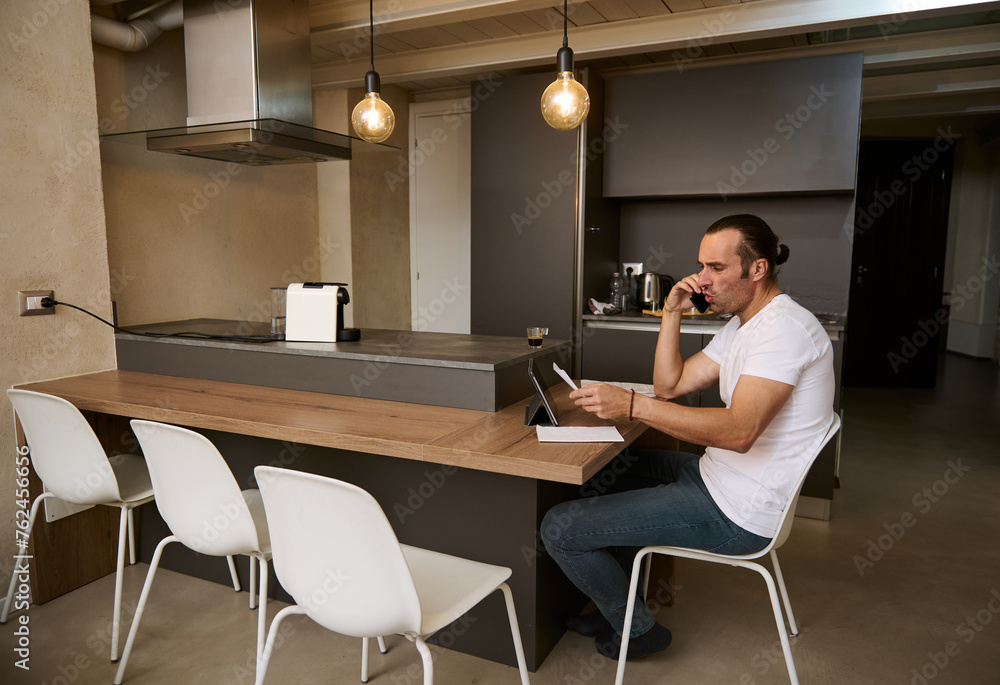 Young man talks on mobile phone, analyzing expenses and costs for paying bills, sitting at desk with documents in hands Young man talks on mobile phone, analyzing expenses and costs for paying bills, sitting at desk with documents in hands