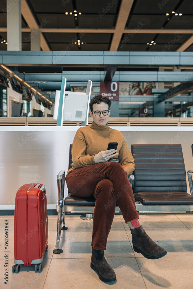 Woman sitting looking at smartphone next to red suitcase in airport lounge