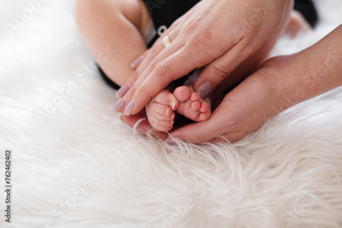 Close-up of baby small legs in moms hands  Ilittle baby dressed in a faithful bodysuit is lying on a white plush blunket