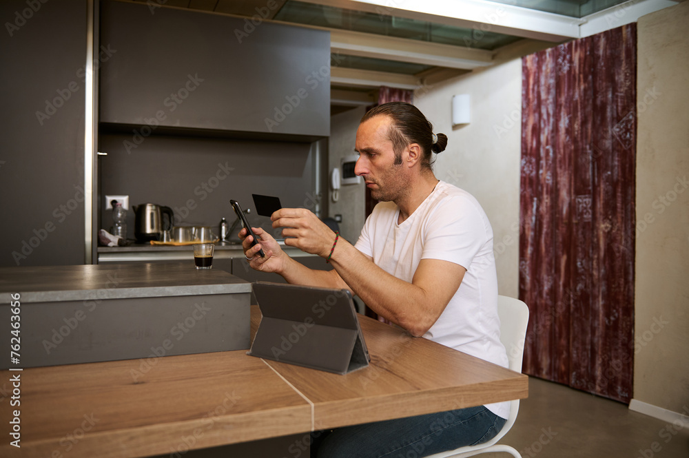 Man making online payments via internet mobile banking, sitting at table with smart phone and credit card in his hands Man making online payments via internet mobile banking, sitting at table with smart phone and credit card in his hands