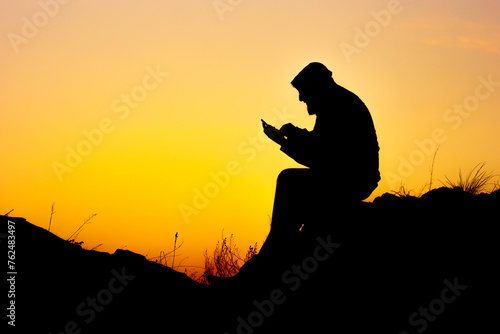 the silhouette of a man sitting on the rocks against the sunset and reading a book