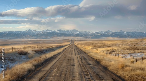 A long dirt road leading through a vast  open prairie towards distant snowy mountains under a dramatic cloudy sky.