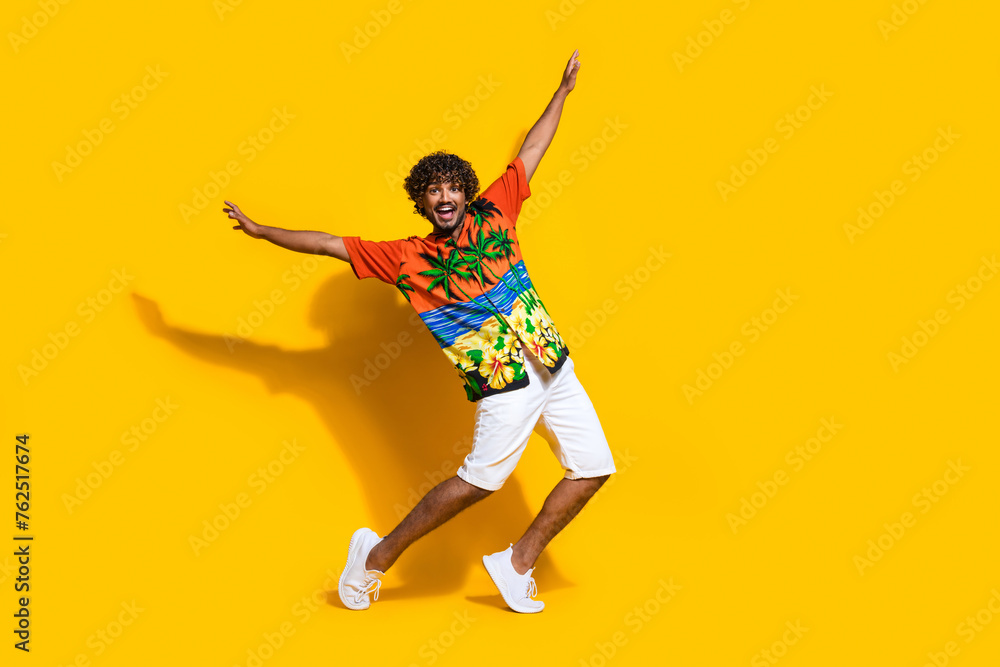Full size photo of ecstatic man dressed print shirt white shorts raising arms dancing on summer party isolated on yellow color background