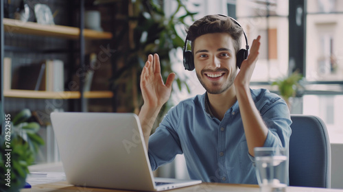 A young businessman in a headset smilingly talks on a video call on a laptop. Sitting in the office at the table and holding a business meeting, training, greeting and waving at the camera photo