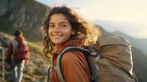 Woman with long hair is smiling and wearing backpack. She is standing on mountain with man behind her