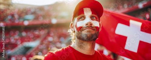Happy Swiss male supporter with face painted in Swiss flag displays a white cross in the centre of a square red field, Swiss male fan at a sports event such as football photo