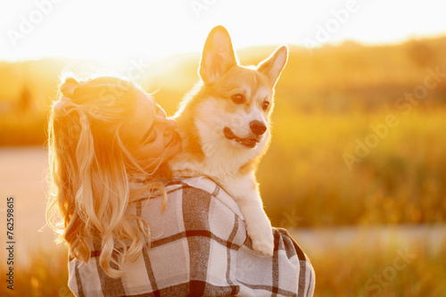 Woman holding Pembroke Welsh Corgi dog during golden summer sunset, hugging tenderly, sharing joy happy moment. Concept of caring for a pet, a walk in nature.