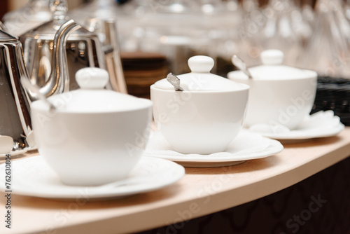 three small white tea cups with lids and spoons on saucers on counter  tableware for cafes  restaurant equipment