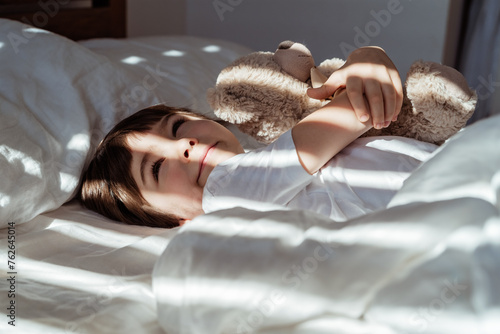Portrait of little happy toddler boy lying in bed hugging his teddy bear plush toy smiling and daydreaming at peaceful sunny morning