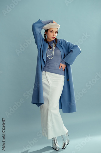 Fashionable confident woman wearing beret, elegant blue coat, turtleneck, layered pearl necklace, white denim maxi skirt, silver boots, posing on blue background. Full-length studio portrait 