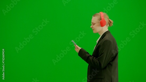 Fototapeta Naklejka Na Ścianę i Meble -  Portrait of female in suit on chroma key green screen. Blonde business woman in formal outfit walking in headphones listening music on smartphone. Side view.