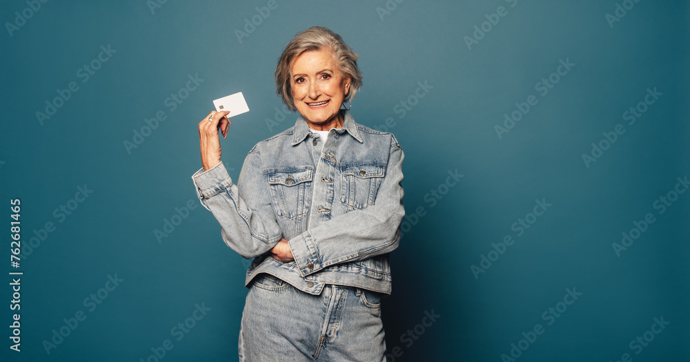 Senior woman with happy smile and credit card standing against blue denim background