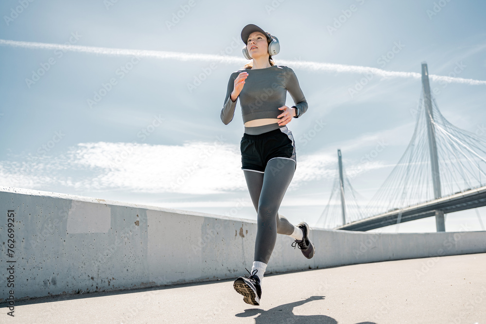 Naklejka premium Dedicated female runner jogging on a bridge with headphones and clear blue skies.