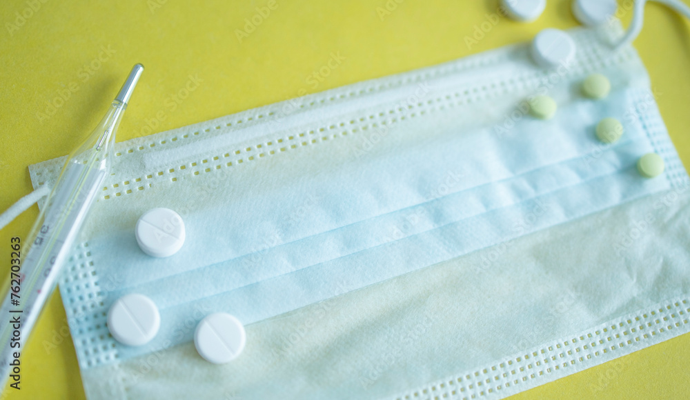 Protective, medical mask, next to the tablets and thermometer and the yellow background. Season of colds and viruses, infections protection Protective, medical mask, next to the tablets and thermometer and the yellow background. Season of colds and viruses, infections protection