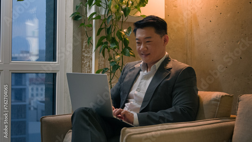 Korean Chinese businessman ethnic Asian man working with laptop at modern office skyscraper male employer business worker CEO manager distant work in chair complete done project close computer smiling