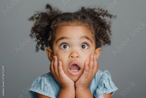 A kind girl with big eyes and a surprised expression on her face. A mixed-race little girl  expressing surprise  with her mouth open and hands on her cheeks in a studio  with a plain grey background