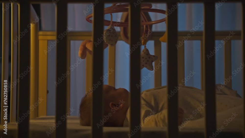 Small child lies in a crib at night getting ready for sleep. The moonlight coming through the window paints the atmosphere of deep night. A night lamp illuminates a small baby in a crib