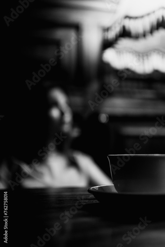 A cup of coffee standing on a wooden table illuminated by warm yellow light in a cozy jazz cafe and a mysterious girl on a background. Black and white.