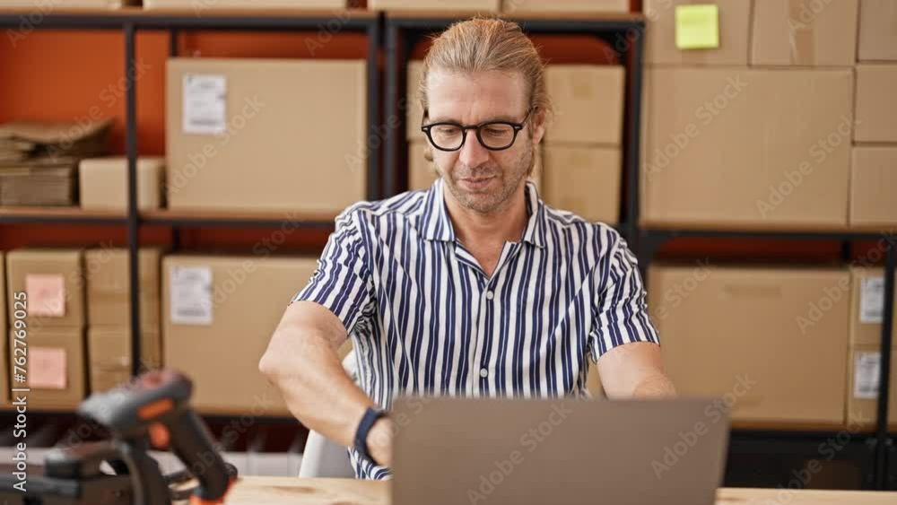 Focused man in glasses working with laptop in warehouse, surrounded by boxes. Focused man in glasses working with laptop in warehouse, surrounded by boxes.