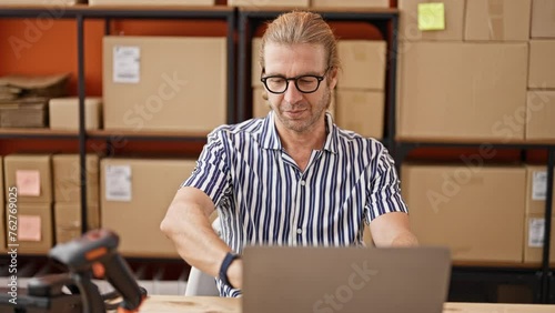 Focused man in glasses working with laptop in warehouse, surrounded by boxes. photo