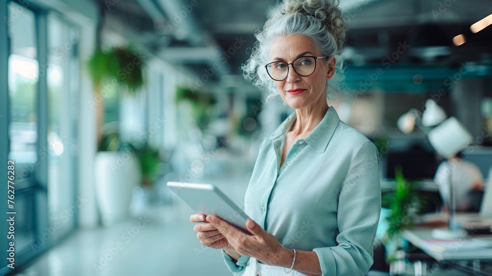 An office worker in her late thirties, with curly gray hair and glasses, reads from a tablet at her desk, dressed in light blue and white business casual. She smiles slightly in a bright, modern corp
An office worker in her late thirties, with curly gray hair and glasses, reads from a tablet at her desk, dressed in light blue and white business casual. She smiles slightly in a bright, modern corp