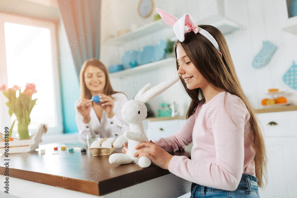 Obraz premium Attractive young woman with little cute girl and boy are preparing for Easter celebration. Happy family wearing bunny ears are spending time together before Easter while painting eggs
