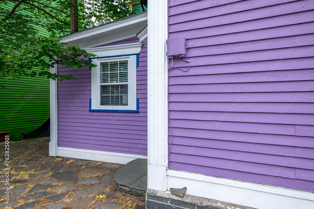 The exterior of a vibrant purple colored wooden wall is covered in horizontal clapboard siding. The building has white trim with thin blue lines. The green maple tree overhangs the sloping roof.  - obrazy, fototapety, plakaty 