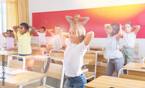 Cheerful preteen pupils doing little warm-up in class with female teacher at lesson in elementary school