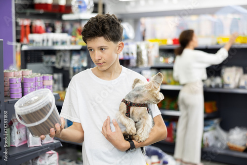 Teenage boy with puppy Yorkshire terrier in arms and carefully read composition of product at packaging of dog dry food, choose healthy food for pet