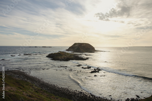 Coastal landscape with a small rocky island surrounded by ocean waves under a cloudy sky at sunset. photo