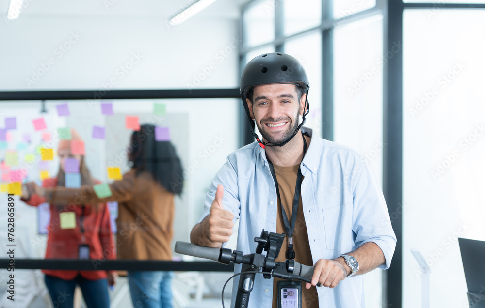 Portrait of smiling businessman riding electric scooter in office with colleague in background Portrait of smiling businessman riding electric scooter in office with colleague in background