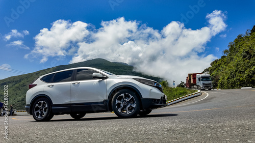 Modern white SUV driving on a scenic mountain road with lush greenery and blue sky with clouds, creating ample copy space on the sky and road surface © fotoworld