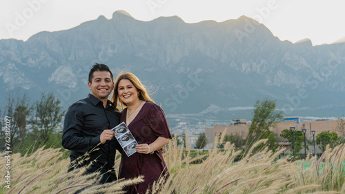 young hispanic couple smiling, holding ultrasound, hill background looking camera