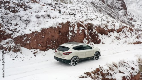 A white car drives along a snow-covered mountain road past red slopes. Konorchek Canyon. Traveling by SUV in Kyrgyzstan. Beautiful mountain winter landscape. photo