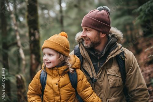 Child in a yellow jacket and beanie smiles beside a bearded man in a forest, both wearing backpacks for hike. Little explorer with bright eyes, clothed in winter gear, shares moment with male guardian