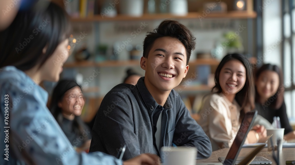 Fototapeta premium Cheerful positive young Asian business professional laughing on corporate meeting with colleagues, sitting at large table, looking away. Diverse team engaged in brainstorming on funny creative ideas