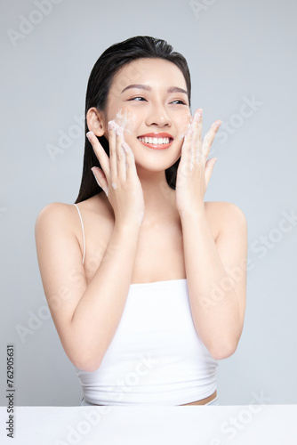 Portrait of cheerful laughing Asian woman applying foam for washing on her face with attractive appearance. Skincare spa relax concept. Isolated on grey background