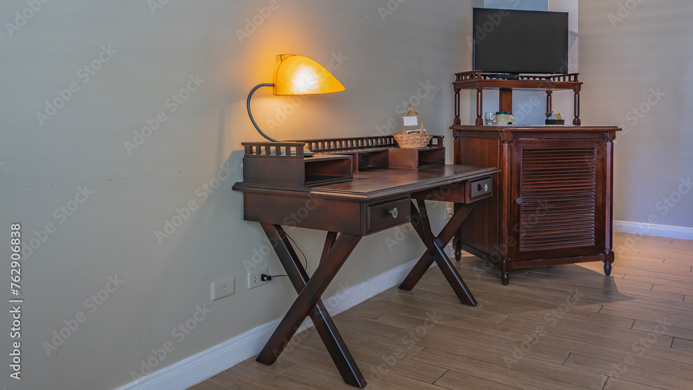 Details of the living room interior in retro style. There is mahogany furniture against the wall. There is a tea set on the chest, a TV. There is a lighted lamp on the table, a wicker basket. Details of the living room interior in retro style. There is mahogany furniture against the wall. There is a tea set on the chest, a TV. There is a lighted lamp on the table, a wicker basket.
