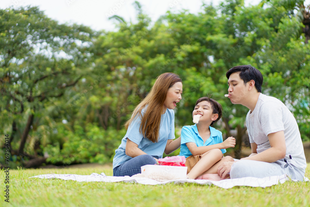 Happy cheerful Asian family with father, mother, and little son enjoy picnic together in a weekend at a park.