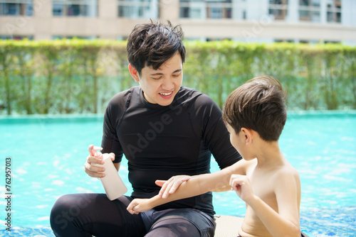 Father is applying a sun screen or sun block lotion on his son body before going to swimming in the swimming pool.  photo