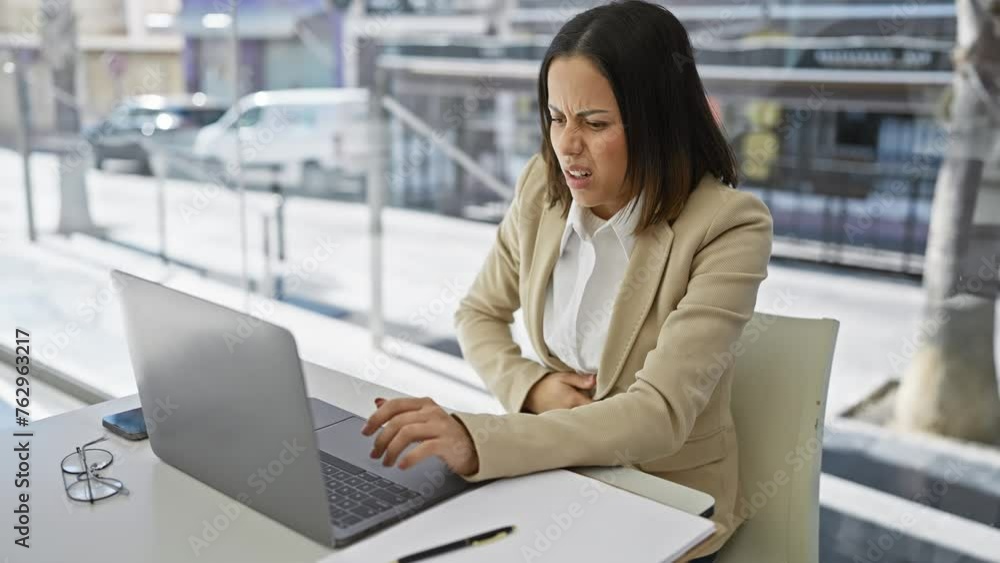 A professional hispanic woman feeling abdominal pain while working on her laptop in a modern office with blurry city background A professional hispanic woman feeling abdominal pain while working on her laptop in a modern office with blurry city background